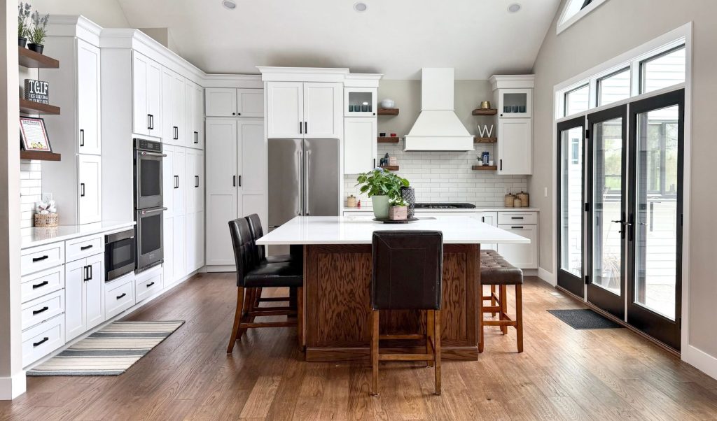 A bright, modern kitchen remodel by Benson Stone Company featuring ivory Marquis Cabinetry, a large wood-toned central island with white countertops, and vaulted ceilings. The space includes integrated stainless steel appliances, dark leather barstools, and large black-trimmed glass doors leading to an outdoor area.