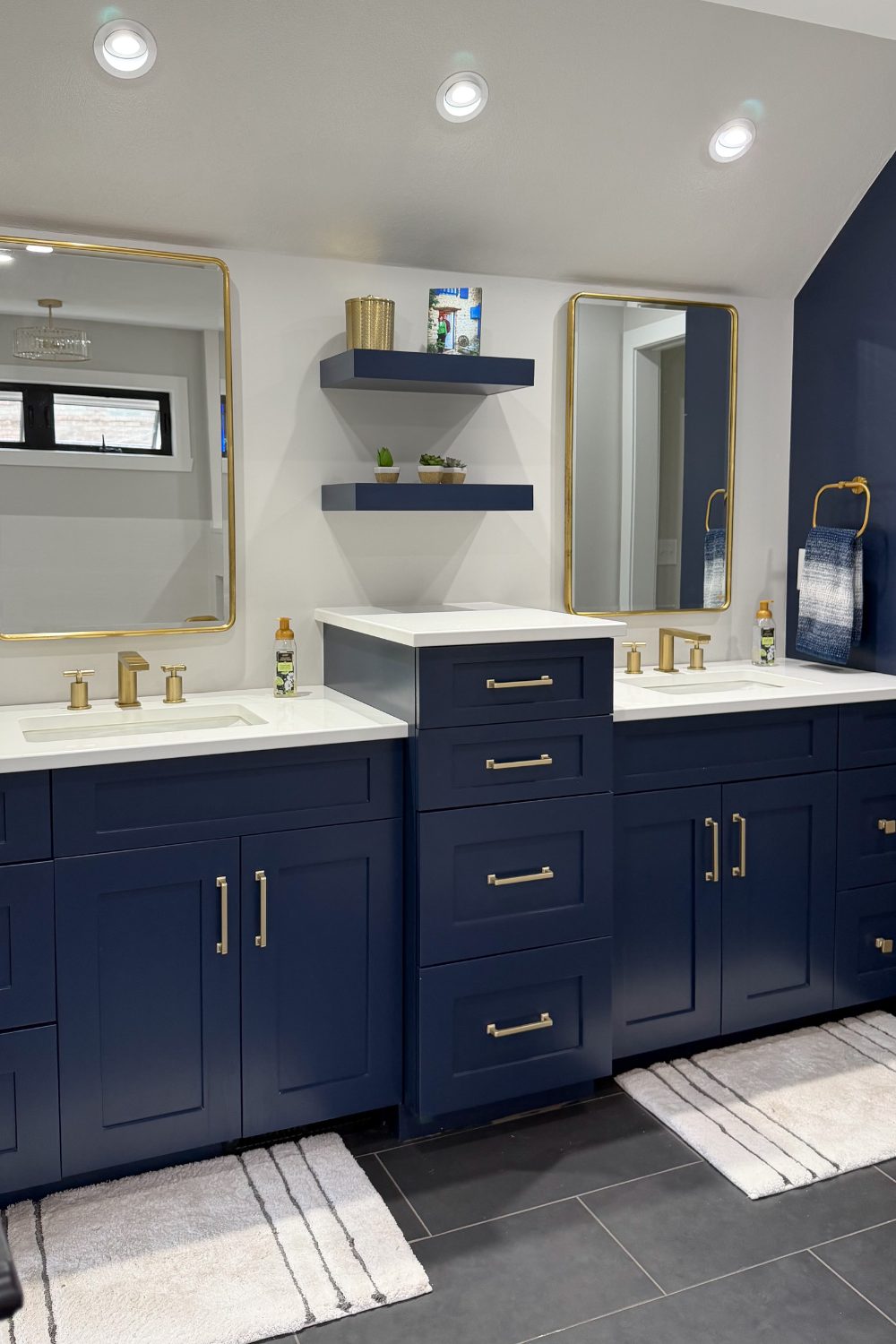 Modern classic bathroom double vanity featuring bold navy blue cabinetry with a stepped-up center tower, clean white countertops, brushed gold faucets, and rectangular gold-rimmed mirrors.