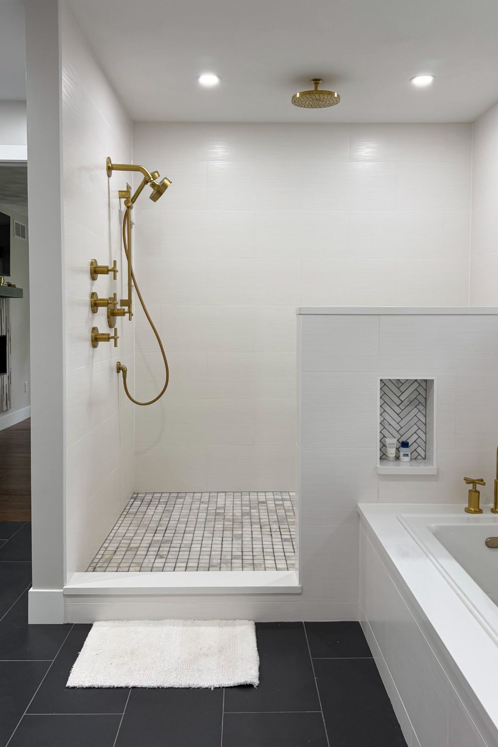 Walk-in master shower with white horizontal-plank wall tiles, curbless entry, grey-and-white mosaic tile floor, and luxurious multi-outlet brushed gold shower fixtures.