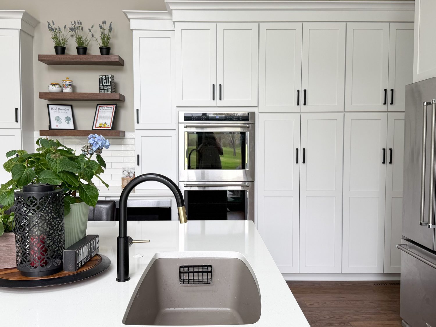 Close-up of a white quartz kitchen island showing a stainless steel undermount sink, a matte black and brass faucet, and a potted hydrangea against a background of white shaker cabinetry.