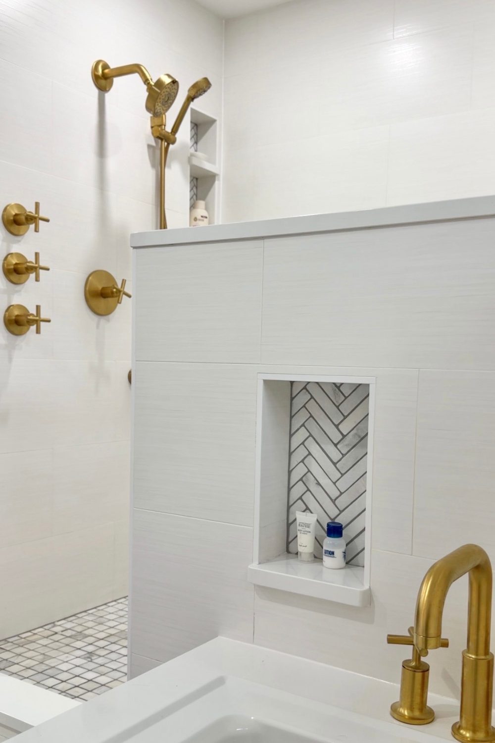 Walk-in master shower with white horizontal-plank wall tiles, curbless entry, grey-and-white mosaic tile floor, and luxurious multi-outlet brushed gold shower fixtures.