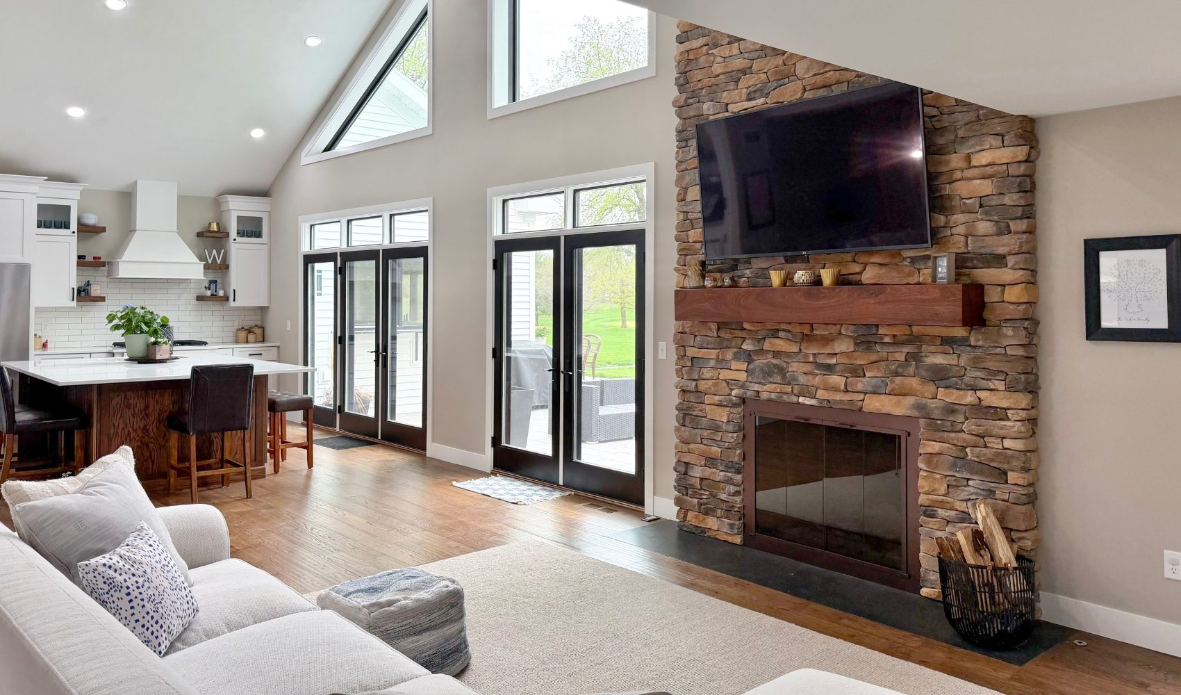An open-concept living room and kitchen remodel by Benson Stone Company featuring a floor-to-ceiling rustic stone fireplace with a thick wood mantel. The "Modern Luxury" space includes vaulted ceilings with recessed lighting, black-framed French doors leading outside, and warm hardwood floors that connect the seating area to the kitchen.