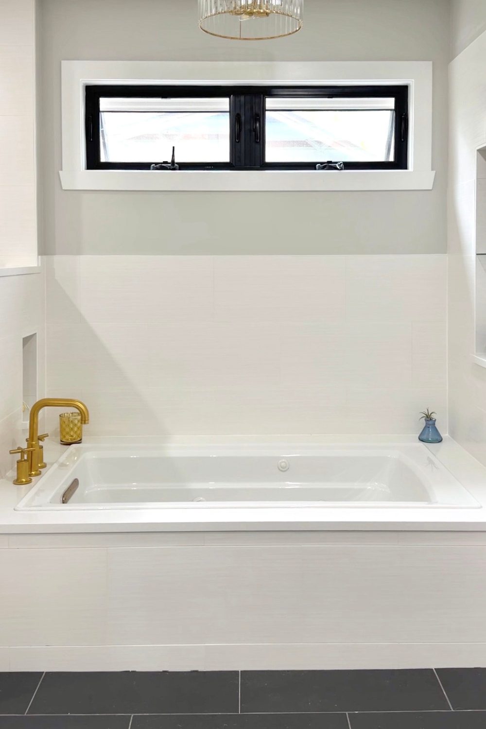 White drop-in soaking tub tucked under a black-trimmed privacy window, flanked by clean white plank wall tiles, black floor tiles, and a contemporary deck-mounted brushed gold faucet.