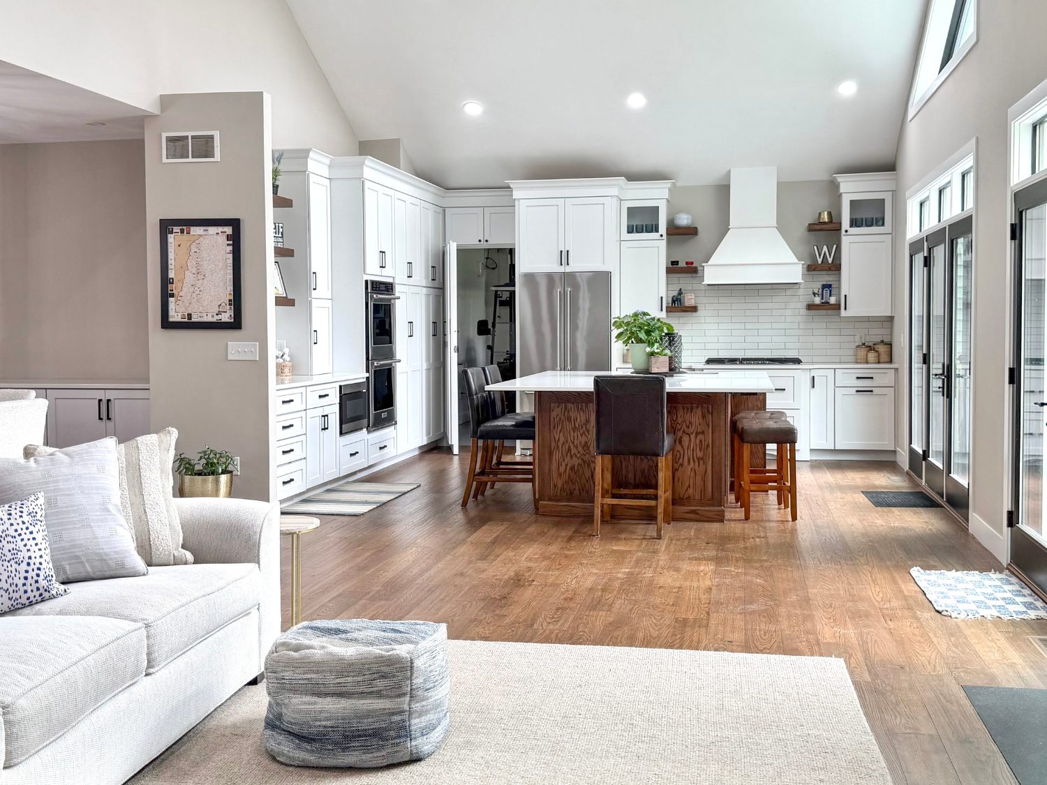 Bright open-concept home layout showing a cozy living room with a white textured sofa, blue accents, and a stone fireplace in the foreground, looking into a modern transitional kitchen.