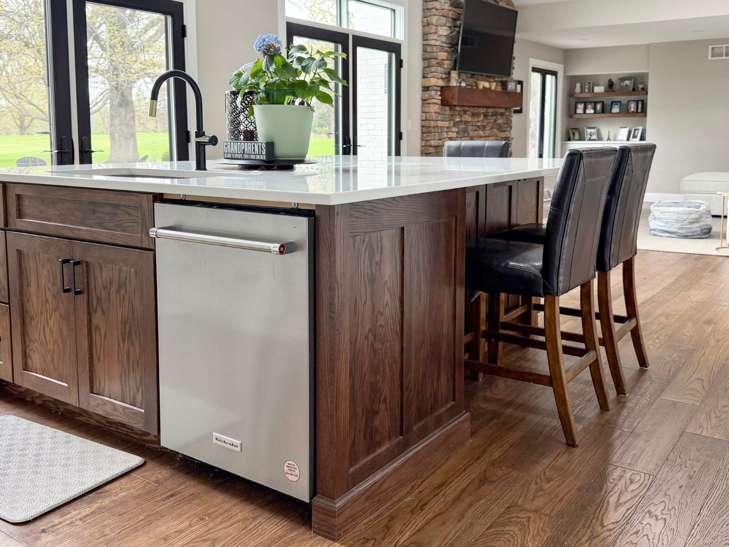 Side profile of a dark wood Maple Dusk kitchen island with white quartz countertops, a built-in stainless steel dishwasher, black leather barstools, and Hallmark Novella Hickory Eliot hardwood floors.
