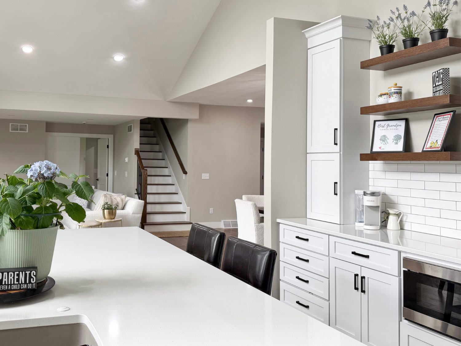 An over-the-sink view of a white quartz kitchen island with a black-and-gold faucet, looking toward custom Birch White shaker cabinets, floating wood shelves, and high-end stainless steel appliances.