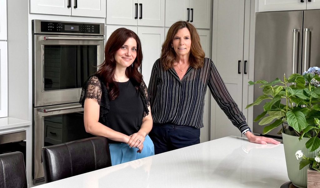 Two women smiling in a professionally designed kitchen at a Benson Stone Company remodel project. The background showcases floor-to-ceiling ivory cabinets with dark hardware, a stainless steel refrigerator, and a sleek white countertop, highlighting the "Modern Luxury" theme of the renovation.