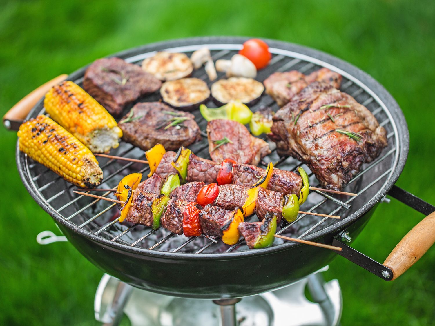 A round kettle grill filled with sizzling food, including beef skewers with bell peppers and tomatoes, corn on the cob, steaks, sliced eggplant, and mushrooms, set against a blurred green grass background.