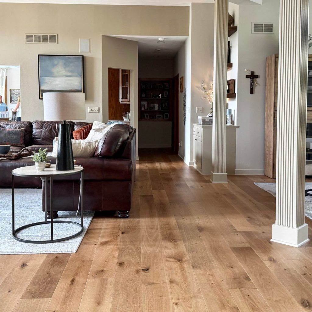 A living room view featuring warm hardwood flooring, a dark leather sectional sofa, and a round side table with a black lamp, looking through to a hallway and built-in shelving.