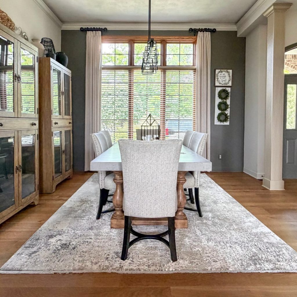 A formal dining room featuring a light-toned concrete-top table with wooden pedestal bases, surrounded by upholstered light gray chairs on a large, textured neutral rug, framed by dark green walls and floor-to-ceiling windows.