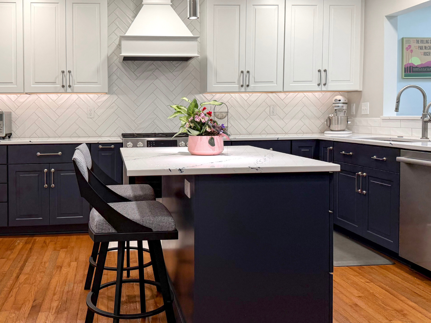 Modern kitchen featuring two-tone navy blue and white cabinetry, a white tile herringbone backsplash, a kitchen island with bar stools, and hardwood floors.